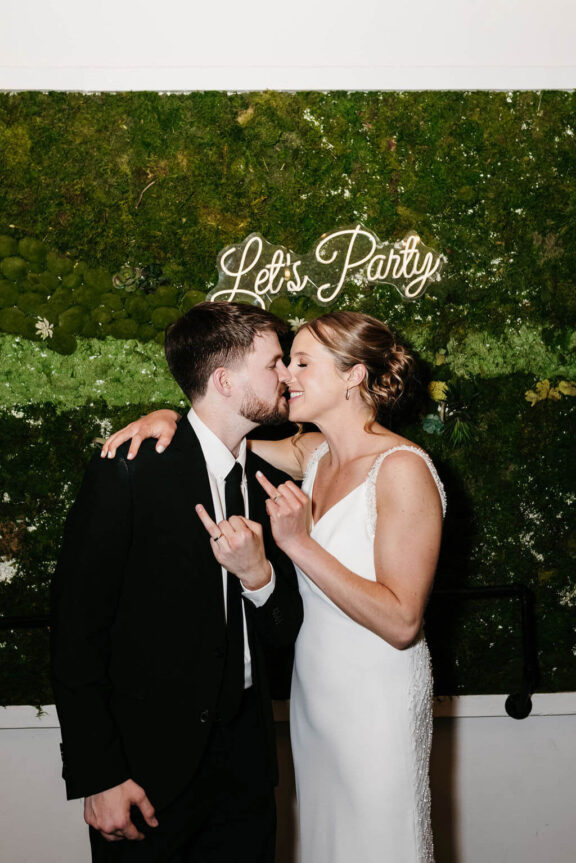 Bride and groom showing off rings in front if a moss wall with a neon light that says "Let's Party"