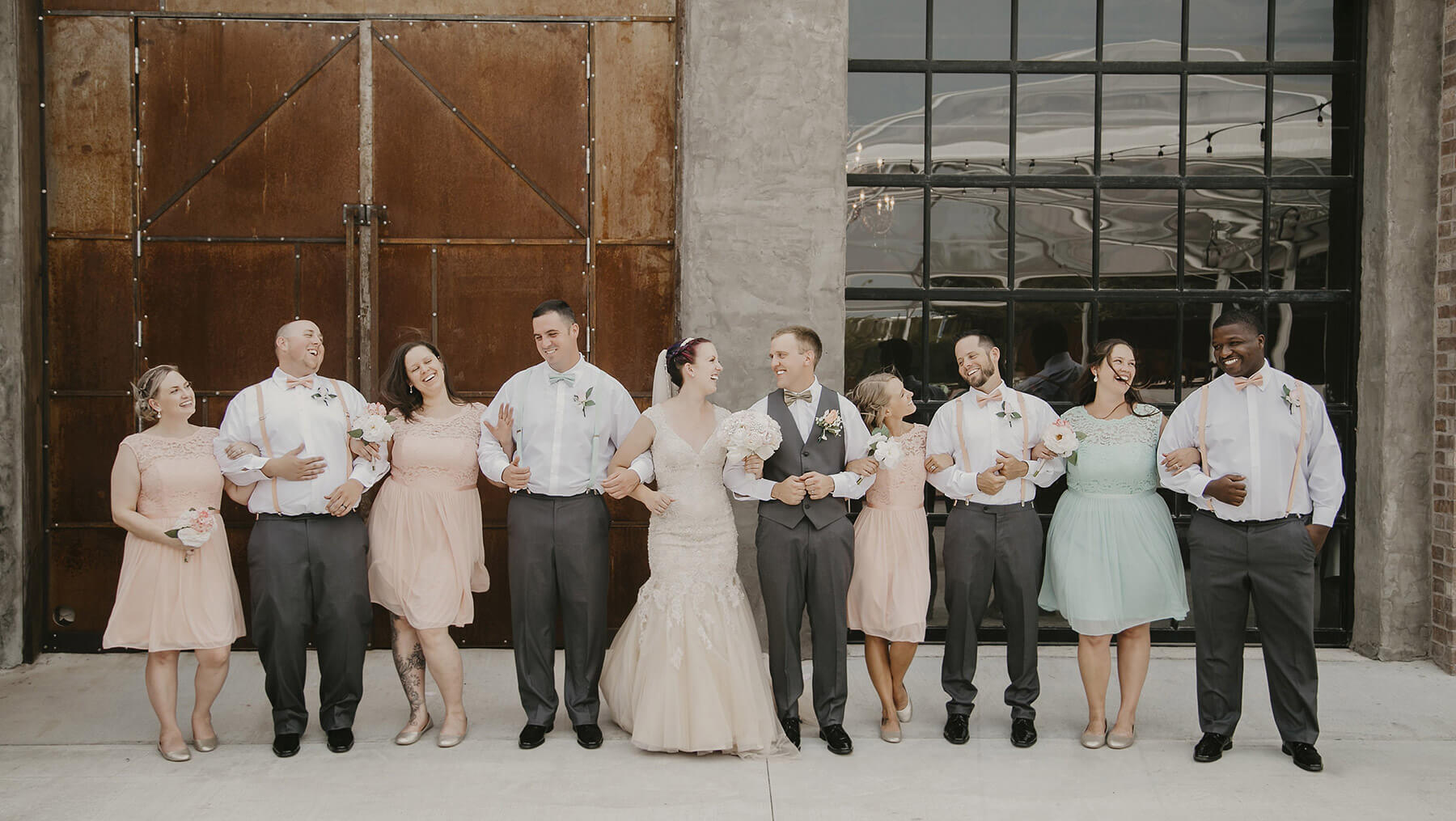 A smiling wedding party linked arm in arm, stands in front of the Brick & Mortar building.